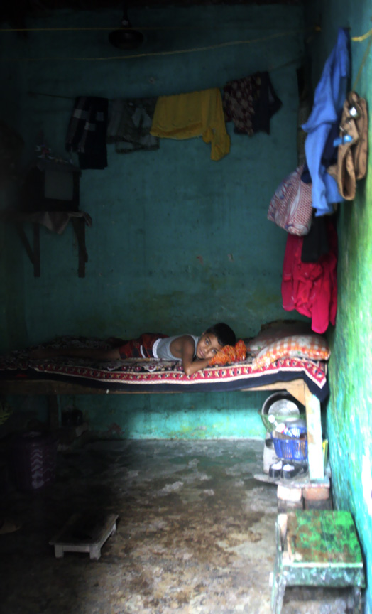 A young boy lies on a bed in a dimly lit room with green walls, surrounded by hanging clothes and household items