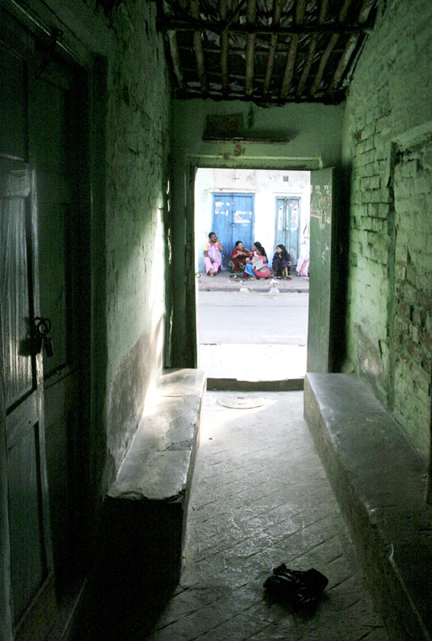 A dimly lit passageway with green walls leads to a sunlit street where a group of women in pink sit and converse near blue doors