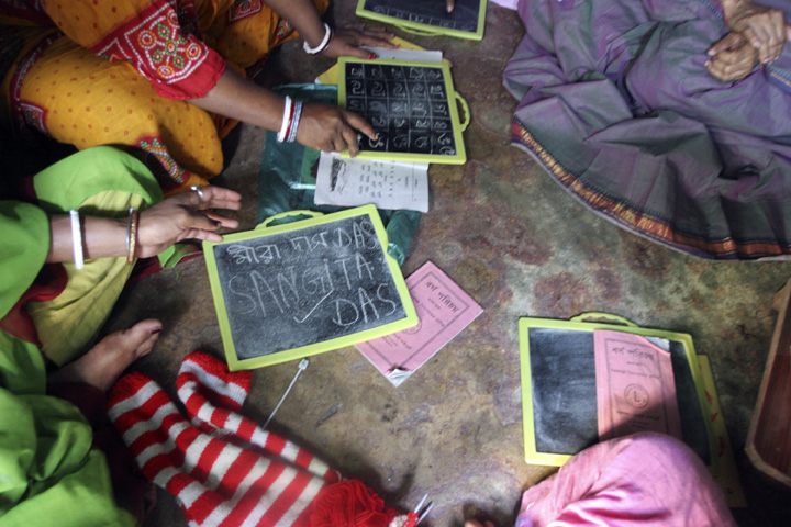 Women sitting on the floor writing on small chalkboards with chalk, participating in a learning session with books and slates