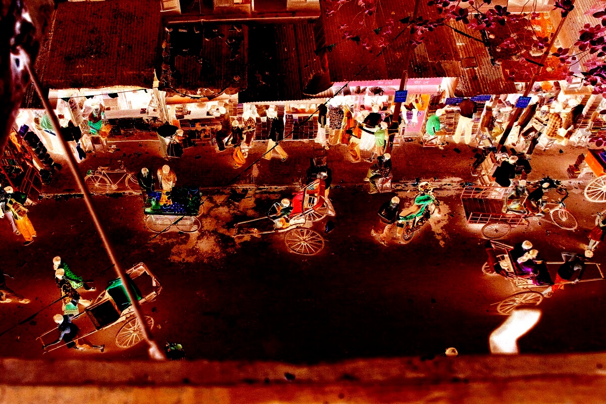 A busy street market with walkers, rickshaws, and vendors selling goods—captured in negative colors