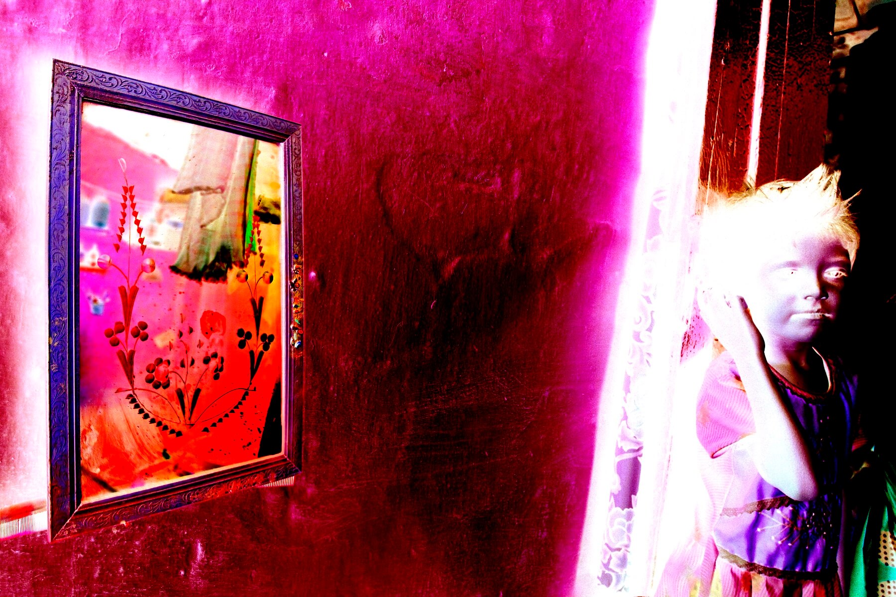 A negative image of a child near a doorway touching their ear; a mirror reflects a patterned curtain and part of a room