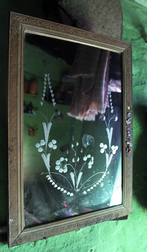 An old mirror with a floral design is mounted on a green wall, reflecting a room with shelves, hanging fabric, and various objects