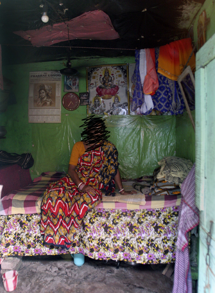 A woman in a patterned sari sits on a bed in a small room with green walls, religious posters, and hanging clothes in the background