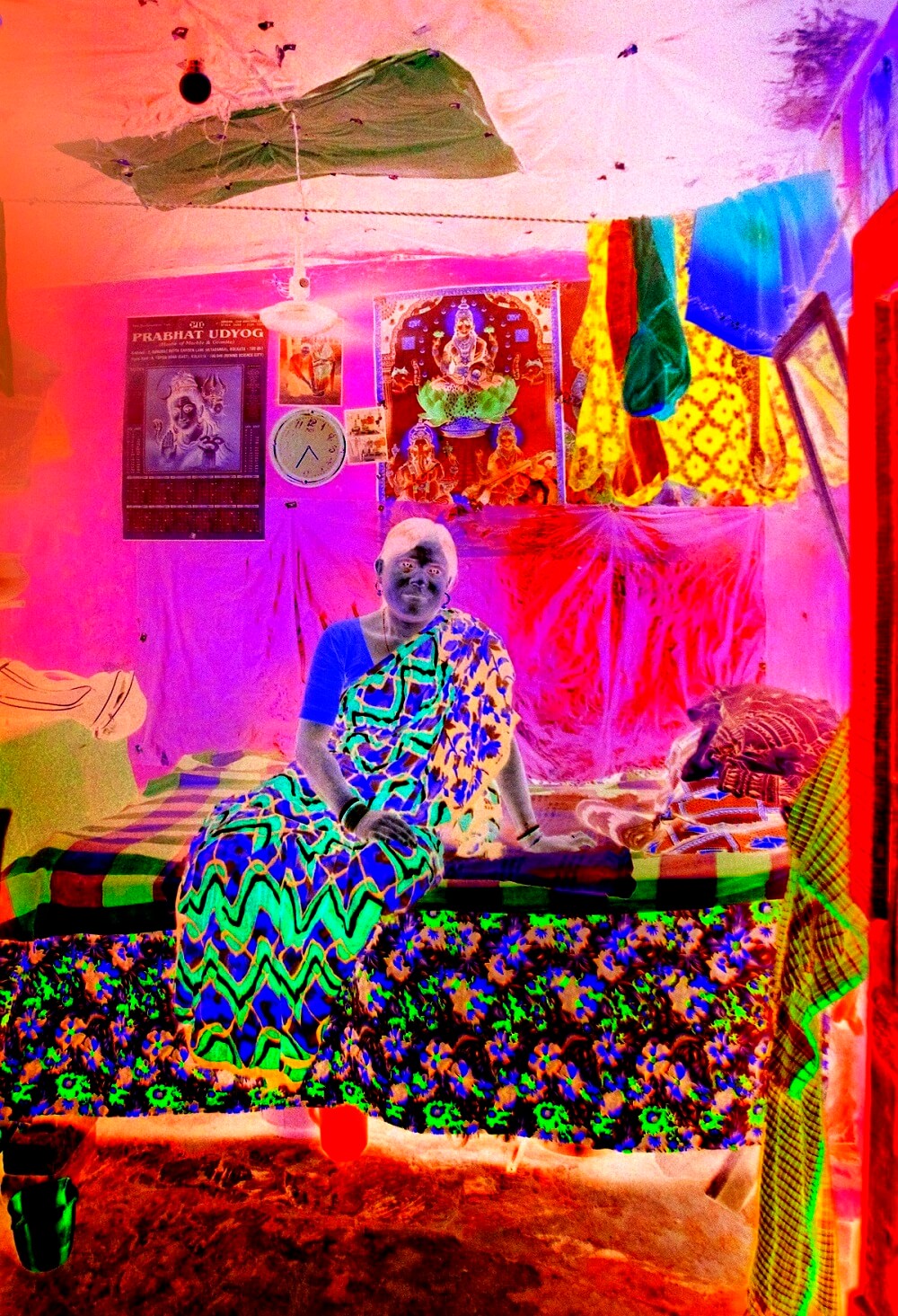 Phool Bibi sits on a bed of God posters, clinging to sanity, with a clock and hanging clothes, looking toward the camera