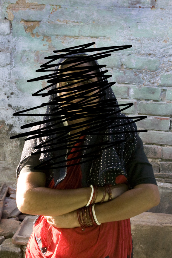 A person wearing a red and black outfit with bangles stands with arms crossed in front of a weathered brick wall. Their face is obscured