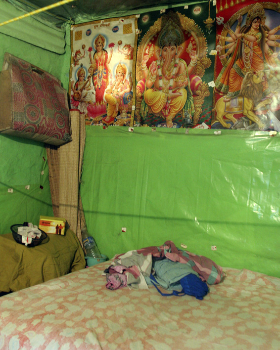 A small room with green plastic-covered walls has religious posters, a bed with folded clothes, and a shelf holding household items