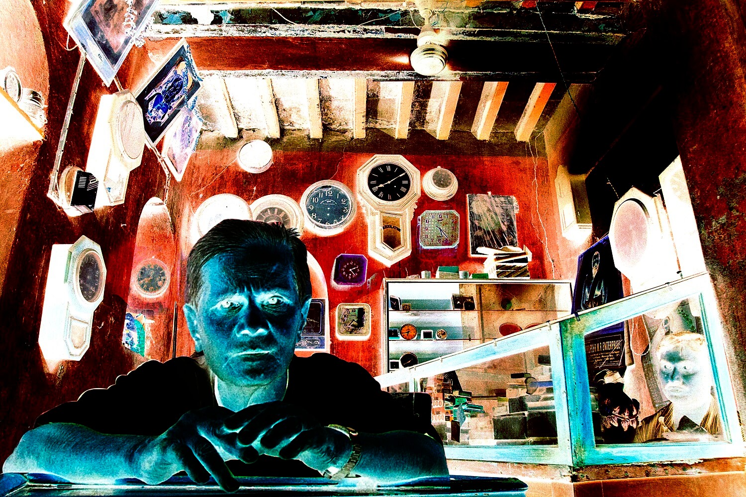 A man sits at a counter in an old clock shop, surrounded by vintage wall clocks—each one frozen in time