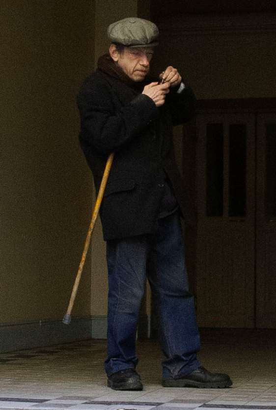 An elderly man in a flat cap and dark coat stands near a building entrance, holding a pipe and using a cane for support