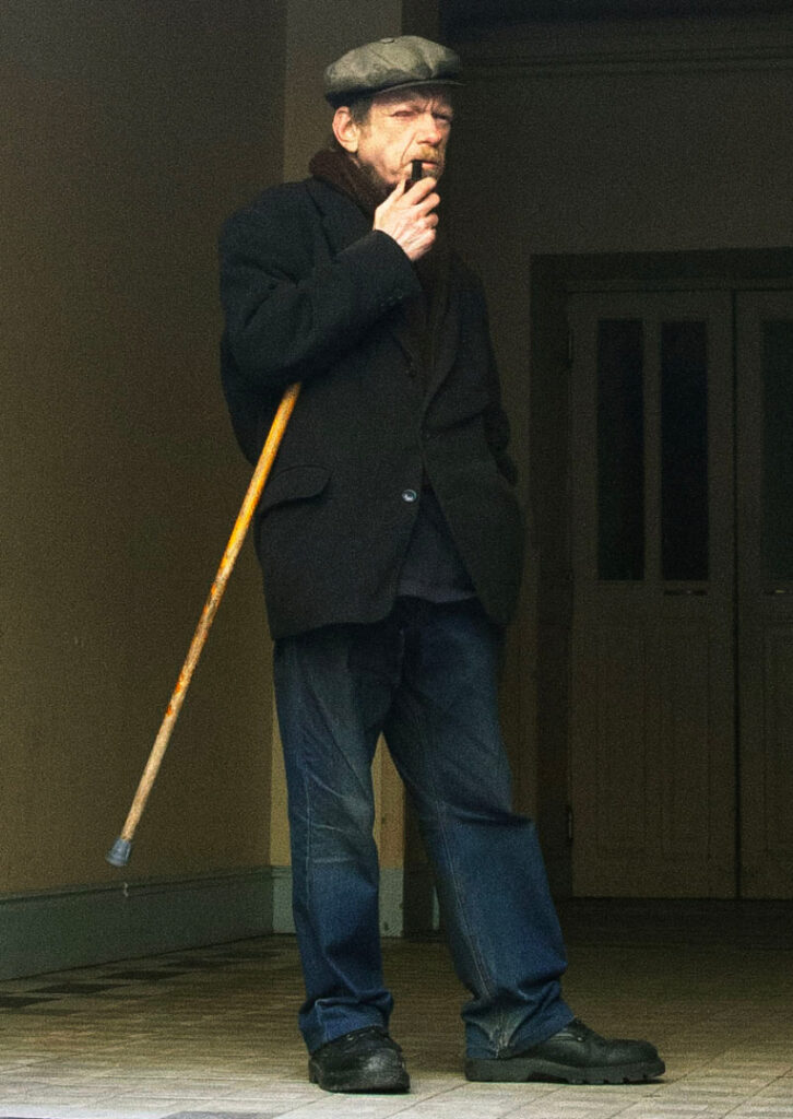 An elderly man in a cap and coat leans on a walking stick, smoking a pipe, standing near a doorway with a thoughtful expression