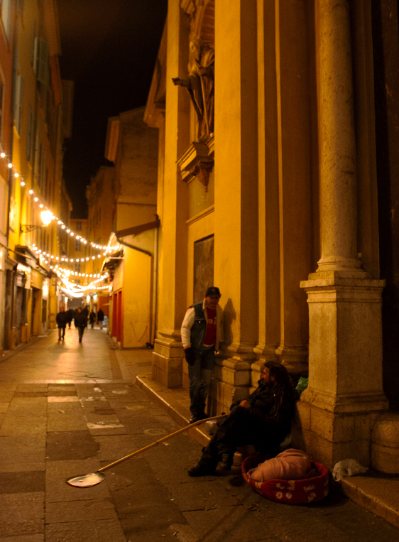 A person sits with a dog on a city sidewalk at night, while another stands nearby. String lights hang above the street