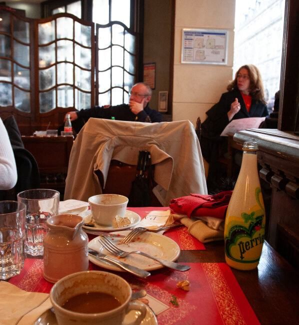 A restaurant table with used dishes, cups, and utensils, with two patrons in the background, one reading a menu and the other seated