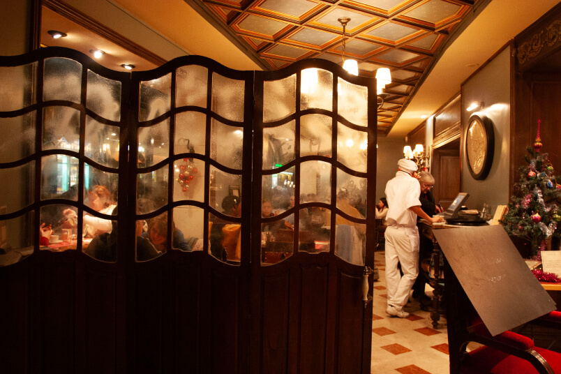 A restaurant with wooden dividers shows diners, a waiter near the counter, and a decorated Christmas tree in the corner