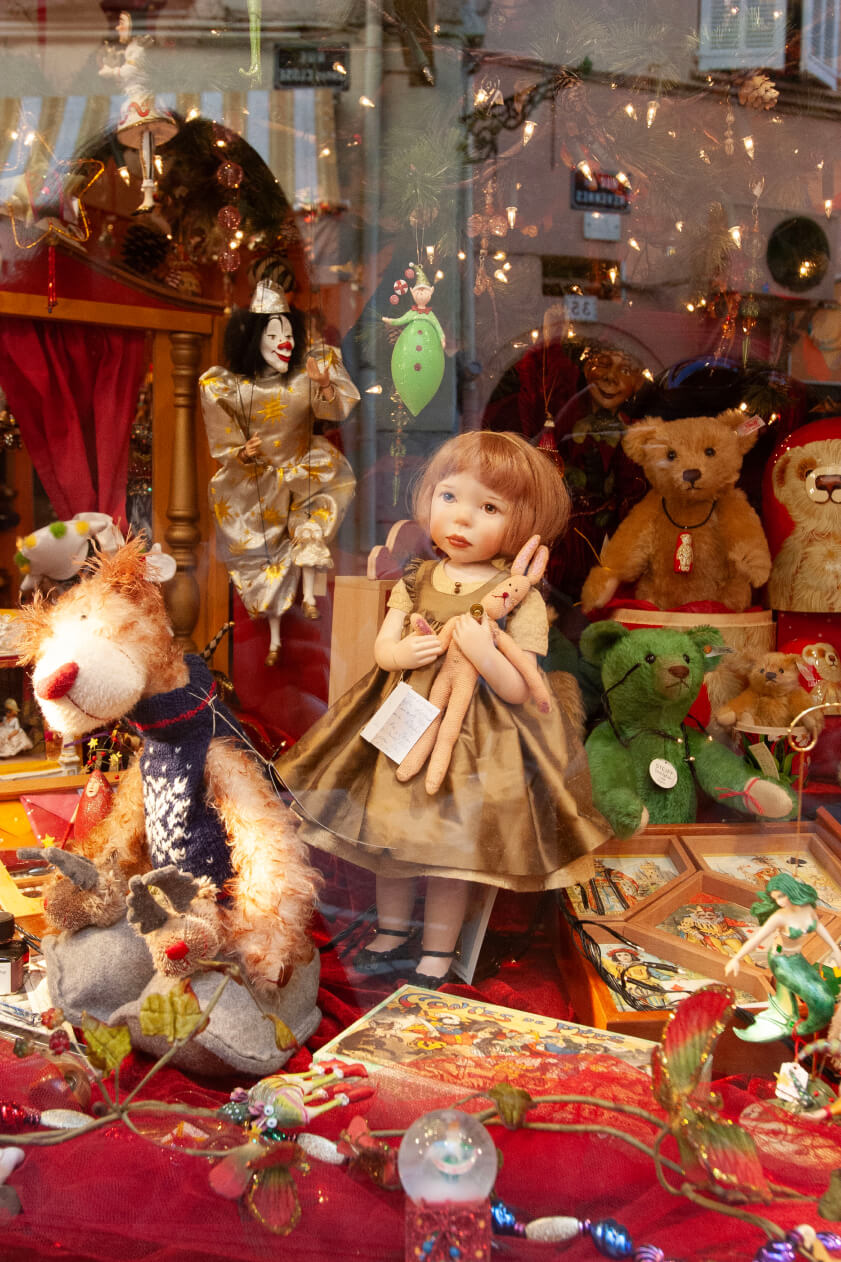 A window display with vintage dolls, stuffed animals, and holiday decorations, reflecting the street through the glass