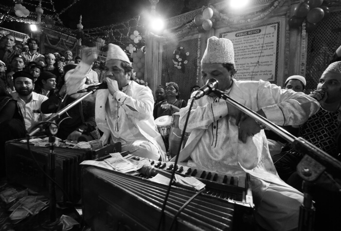 Qawwali singers perform with harmoniums and microphones, musicians accompanying them, as the audience is captivated by the energy