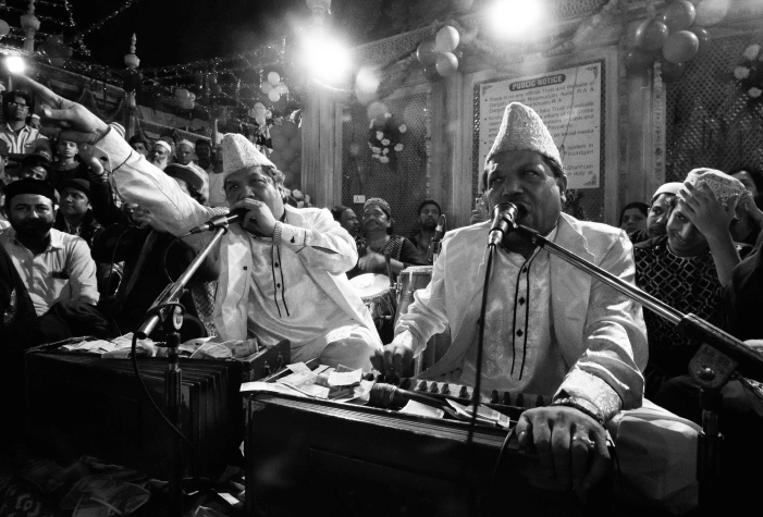 Qawwali performers sing passionately with harmoniums and microphones, surrounded by musicians, drawing in the engaged audience