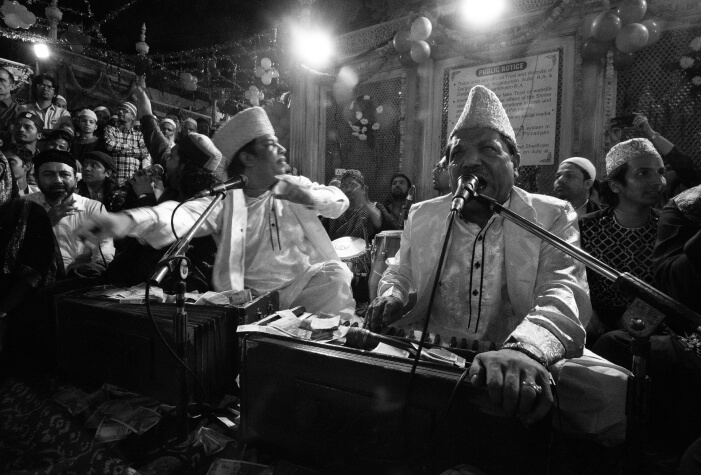 Qawwali singers perform with harmoniums and microphones at a gathering, engaging the audience while musicians accompany them