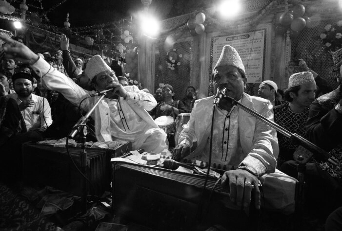 A group of qawwali musicians perform passionately with harmoniums and microphones, surrounded by an engaged audience