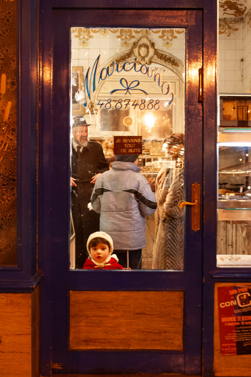 A bakery with a blue door shows people chatting inside, while a child in a red coat gazes out through the glass
