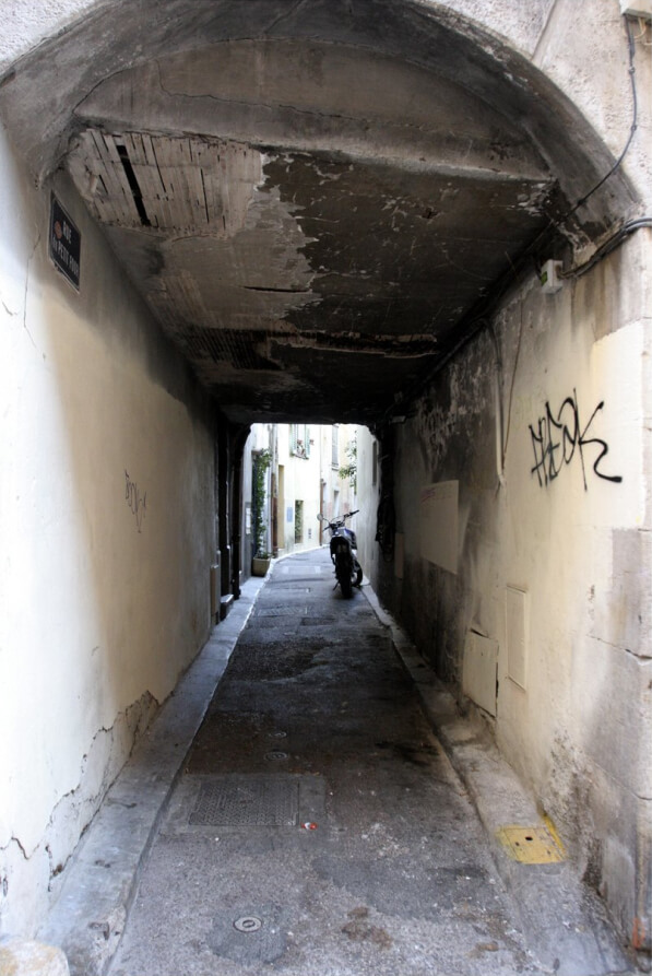 A narrow, arched passage with cracked walls and graffiti leads to a brighter street, where a parked motorcycle is visible