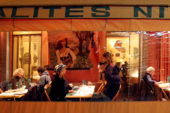 A restaurant with diners seated at tables, a waitress pouring wine, and a painting of a woman with a guitar on the wall inside