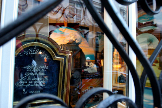 A view through a wrought iron gate into a Parisian shop window, displaying artwork and a sign with cursive script