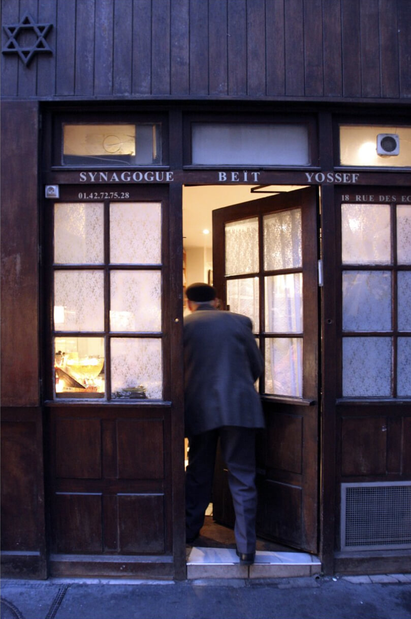 A man in a hat and dark coat enters the wooden doors of Synagogue Beit Yossef, with warm lighting inside