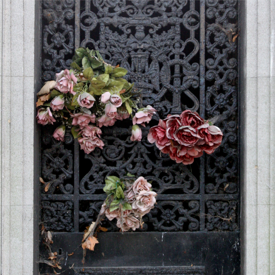 An ornate black metal gate with intricate patterns, featuring faded artificial flowers, some leaves and petals wilting