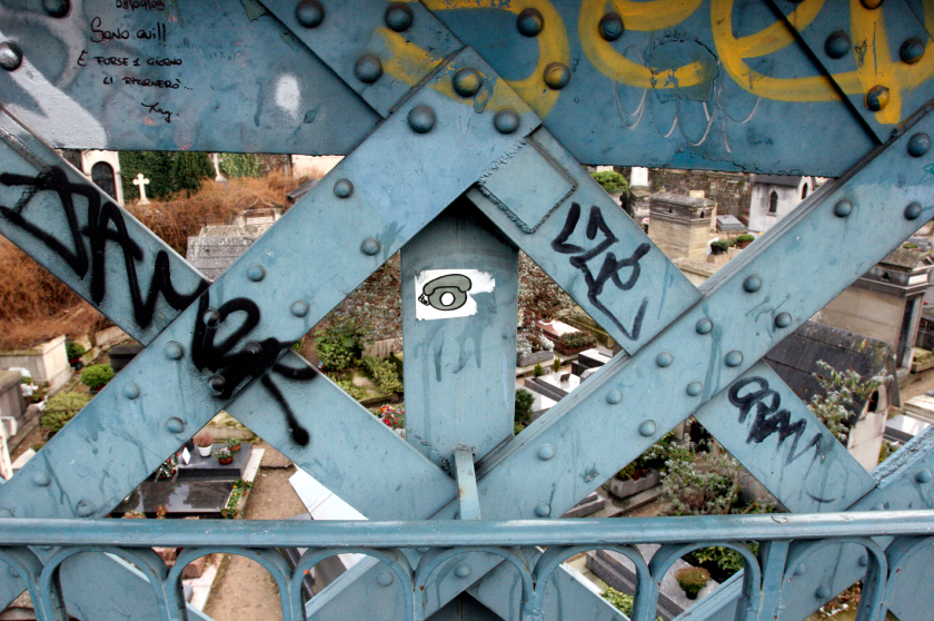 A blue metal bridge with graffiti and a sticker overlooks a cemetery, with tombstones and greenery below