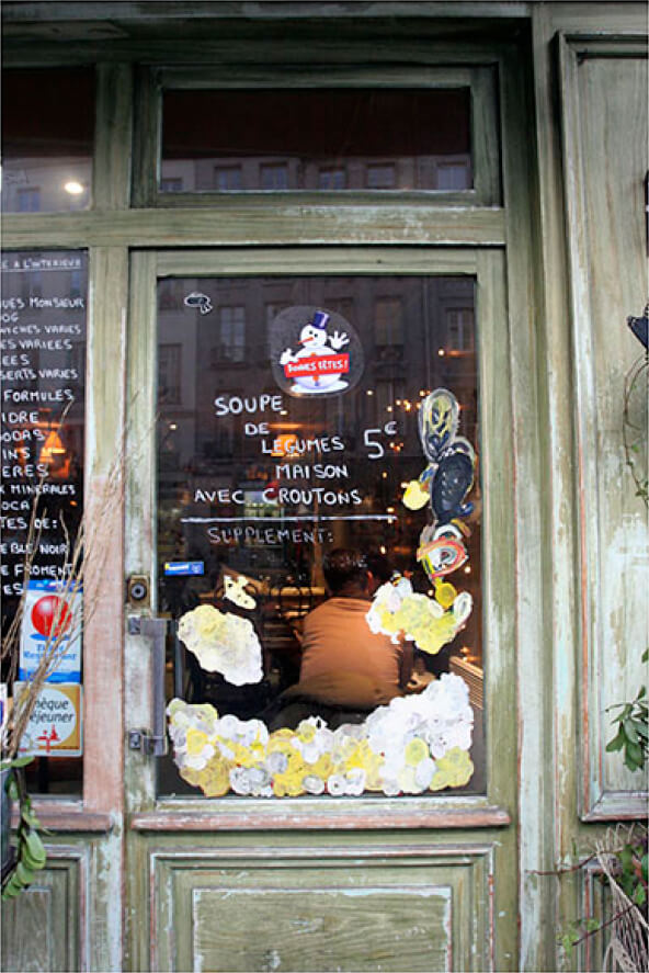 A weathered café door with a festive sticker and handwritten menu reveals a man seated inside, lit by warm, ambient light