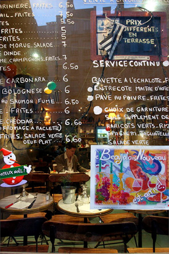 A café window shows a handwritten menu and festive sign, reflecting the street, while a man dines inside