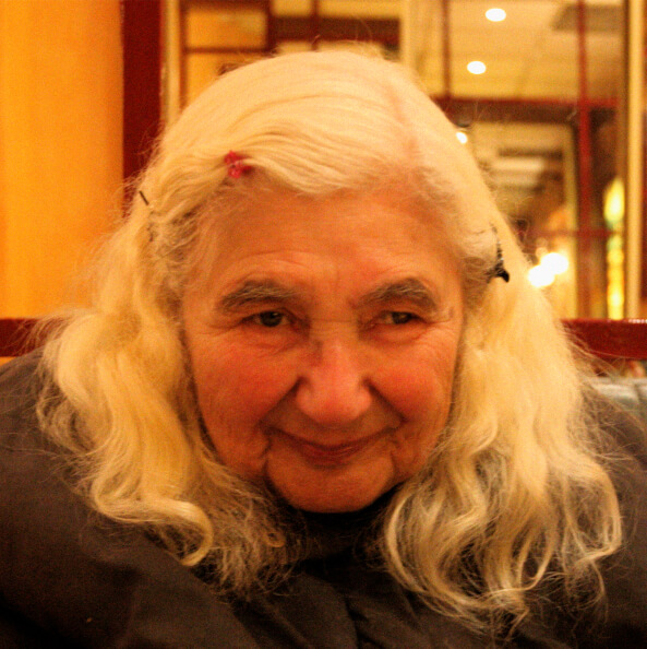 A close-up of an elderly woman with long white hair, wearing hair clips, sitting indoors with a slight smile on her face
