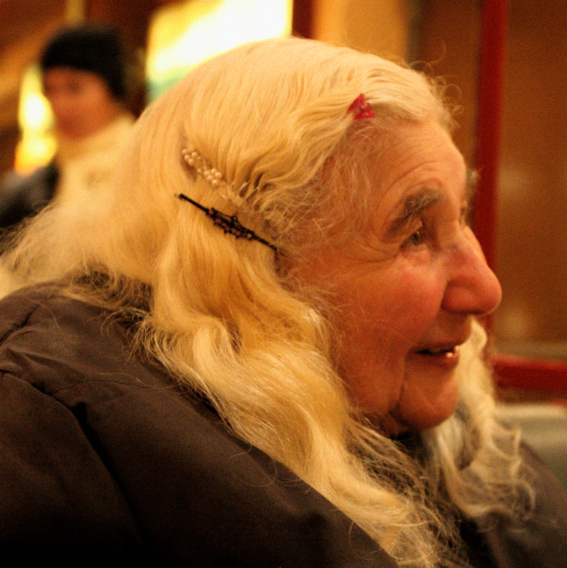 An elderly woman with wavy blonde hair sits indoors, wearing hair clips and a dark coat, smiling as she looks to the side