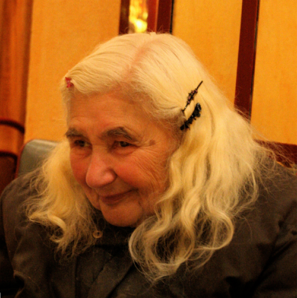 An elderly woman with long white hair, wearing hair clips and a dark coat, sits indoors with a slight smile and a gentle expression