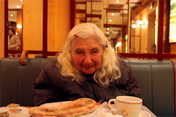 An elderly woman with long white hair sits in a restaurant booth, smiling at the camera, with a meal and tea in front of her