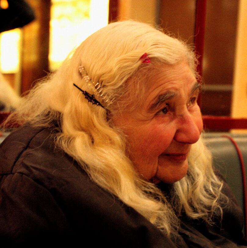 An elderly woman with long, wavy blonde hair sits in a café, wearing hair clips and a dark coat, smiling as she looks to the side
