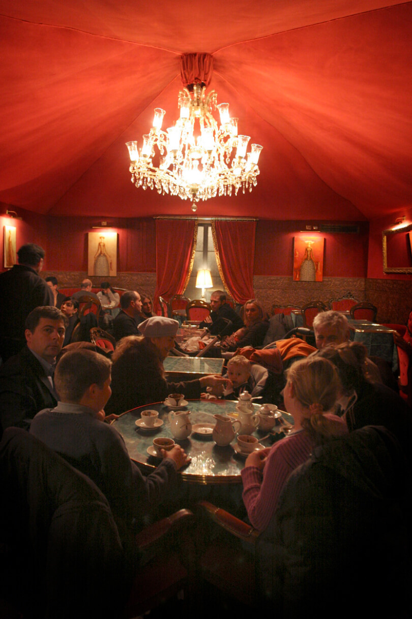 A group of people seated around a table with tea and pastries in a dimly lit room with red walls and a chandelier overhead
