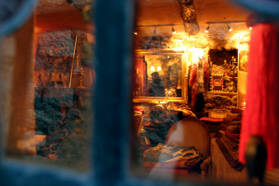 A view through a window into a warmly lit antique store showcasing ornate furniture, mirrors, and vintage textiles