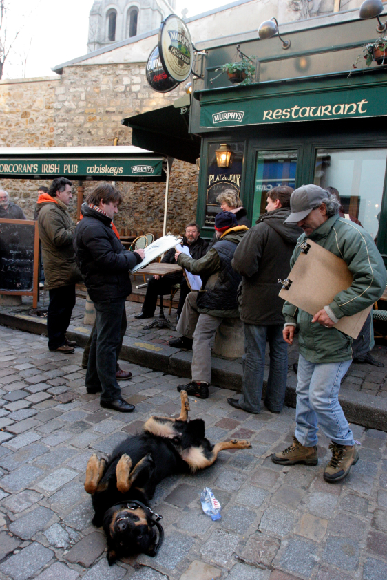 A group of people gathers outside a restaurant, some holding papers, while a large dog lies on its back on the cobblestone street