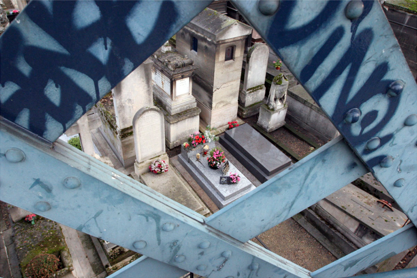 A blue metal bridge with graffiti and a sticker in the foreground overlooks a cemetery, with tombstones and greenery below
