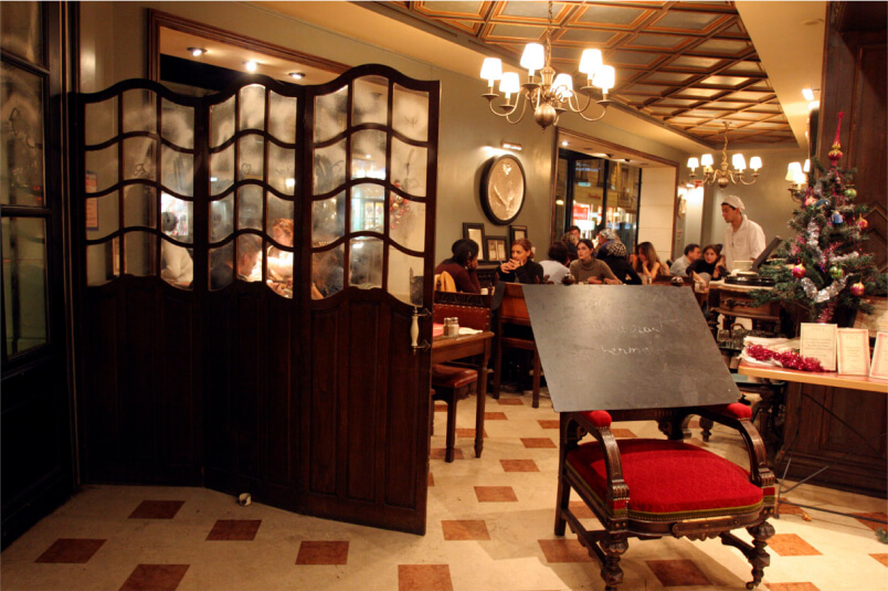 A restaurant interior with diners, a waiter, a wooden divider, chalkboard, and a decorated tree
