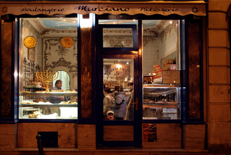 "Murciano Patisserie" glows at night as people browse inside; a child stands at the glass door, looking out