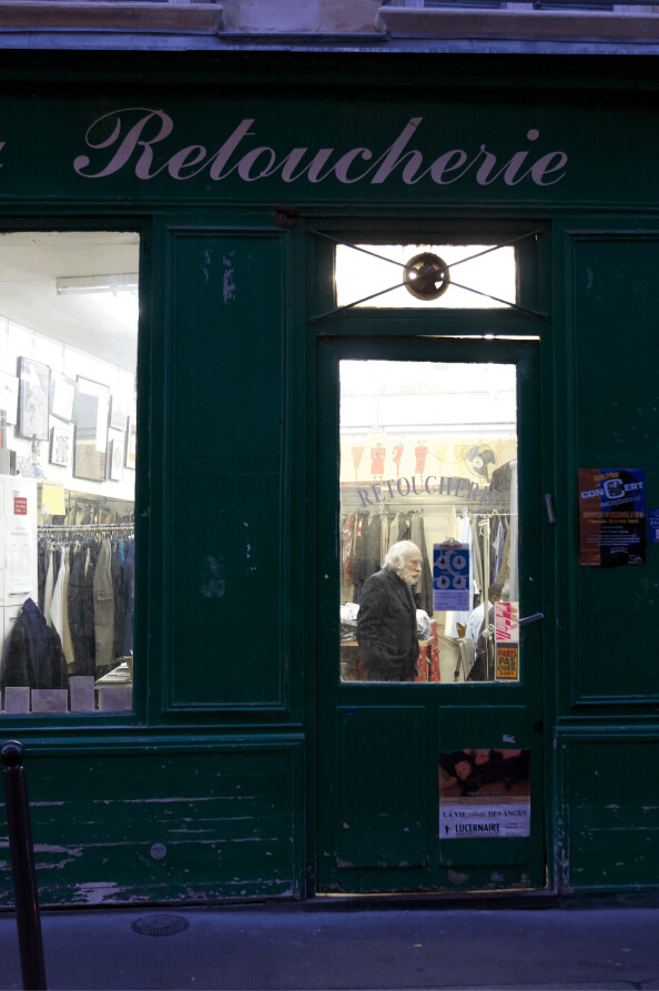 A tailoring shop with a green facade labeled "Retoucherie" shows an elderly man working among clothing racks and garments