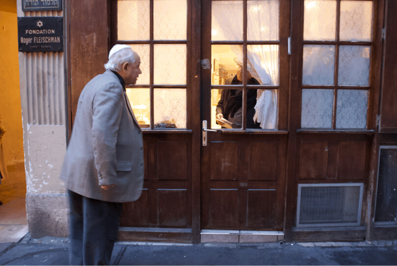 An older man in a gray jacket stands outside a wooden-framed glass door, looking at a warmly lit room with another person inside